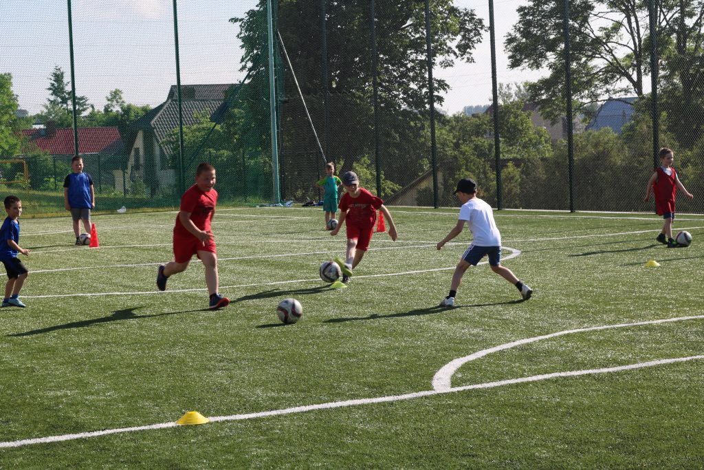 Dwóch chłopców w czerwonych, krótkich strojach sportowych kopie piłki. Za chłopcami, po lewej stronie dwóch graczy w niebieskich koszulkach – jeden z nich stoi przy czerwonym plastikowym pachołku i ma przy nogach piłkę. Natomiast za chłopcami po prawej stronie stoi gracz w jasnozielonym stroju, również z piłką przy nogach. Na wprost chłopców w czerwonych strojach biegnie odwrócony tyłem zawodnik w białej koszulce i ciemnej czapce z daszkiem. Obok niego z piłką stoi chłopiec w czerwonej koszulce. Między zawodnikami na murawie rozrzucone są plastikowe pachołki koloru żółtego. W oddali za długą zieloną siatką ogradzającą boisko widoczne są zielone drzewa i dachy domów. 
