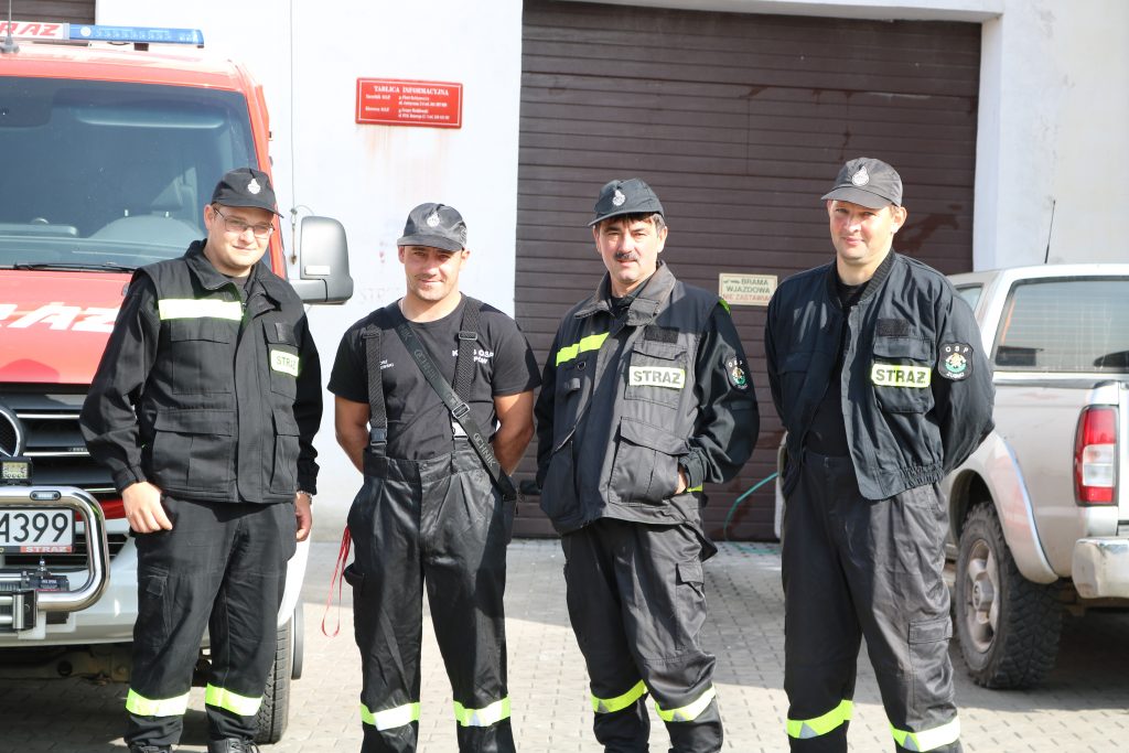 Na zdjęciu czterech strażaków z OSP Gminy Filipów w czarnych strojach strażackich z czapkami z daszkiem. Za strażakami widoczny czerwony samochód strażacki 
