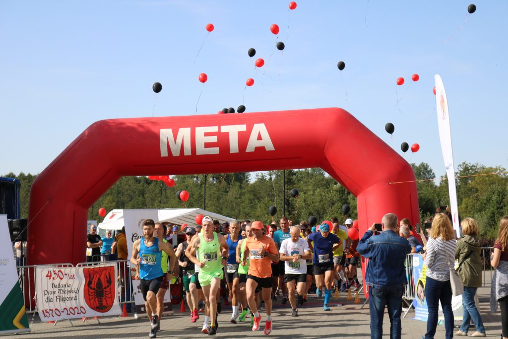 Na zdjęciu tłum biegaczy w kolorowych strojach sportowych wybiegający spod czerwonej, pompowanej bramy startowej. Nad bramą, na błękitnym niebie unoszą się czerwone i czarne balony. W tle zielone drzewa rosnące za placem. 
