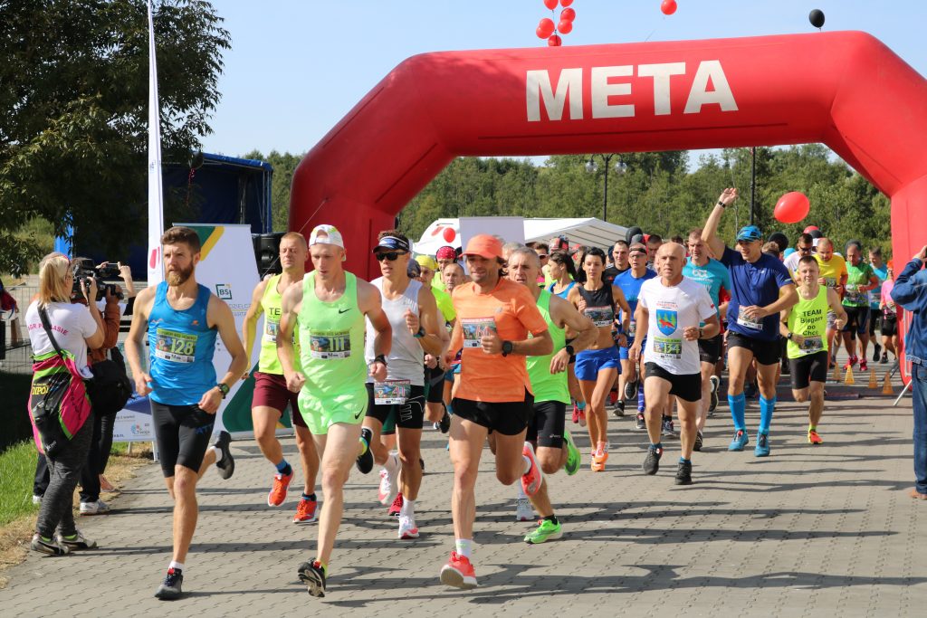 Na zdjęciu tłum biegaczy w kolorowych strojach biegnący w peletonie. Za nimi duża, czerwona, pompowana brama startowa z napisem META.