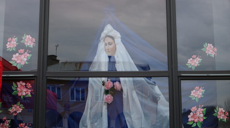Zdjęcie przedstawia umieszczoną za witryną budynku GOKiS ozdobę Matki Boskiej wykonaną z niebieskiego i białego płótna, ozdobioną różowymi kwiatami.