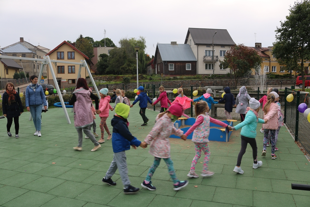 Zdjęcie przedstawia grupę kilkunastu dzieci idących wkoło placu zabaw. Dzieci idą wężykiem i trzymają się za ręce. Na siatce ogradzającej plac zabaw wiszą kolorowe baloniki 