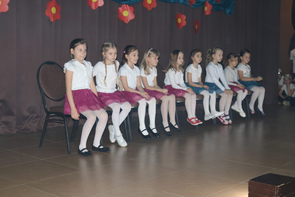 Dziewięć dziewczynek ubranych w białe rajstopki i bluzeczki oraz krótkie czerwone i niebieskie spódniczki. Dziewczynki siedzą na krzesełkach na scenie i za chwilę zaczną swój układ taneczny.