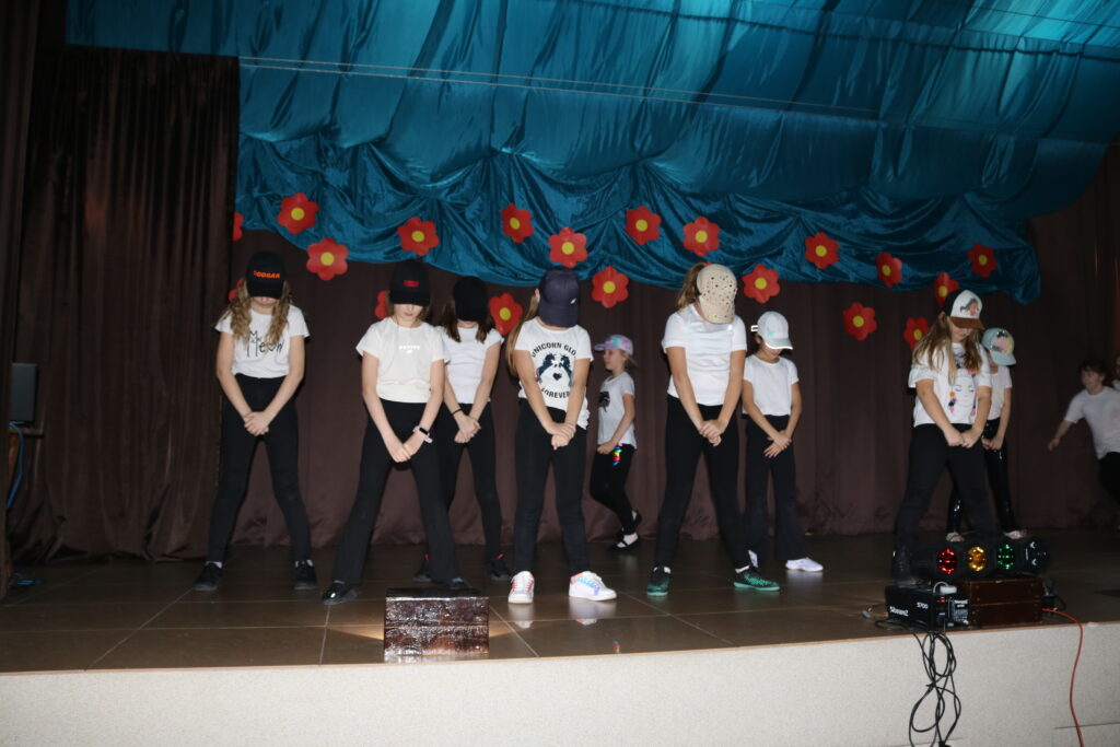 Na scenie grupa HIP HOP w swoim pokazowym wykonaniu. Dziewczyny ubrane w czarne spodnie i białe bluzki, a na głowach czapeczki z daszkiem.