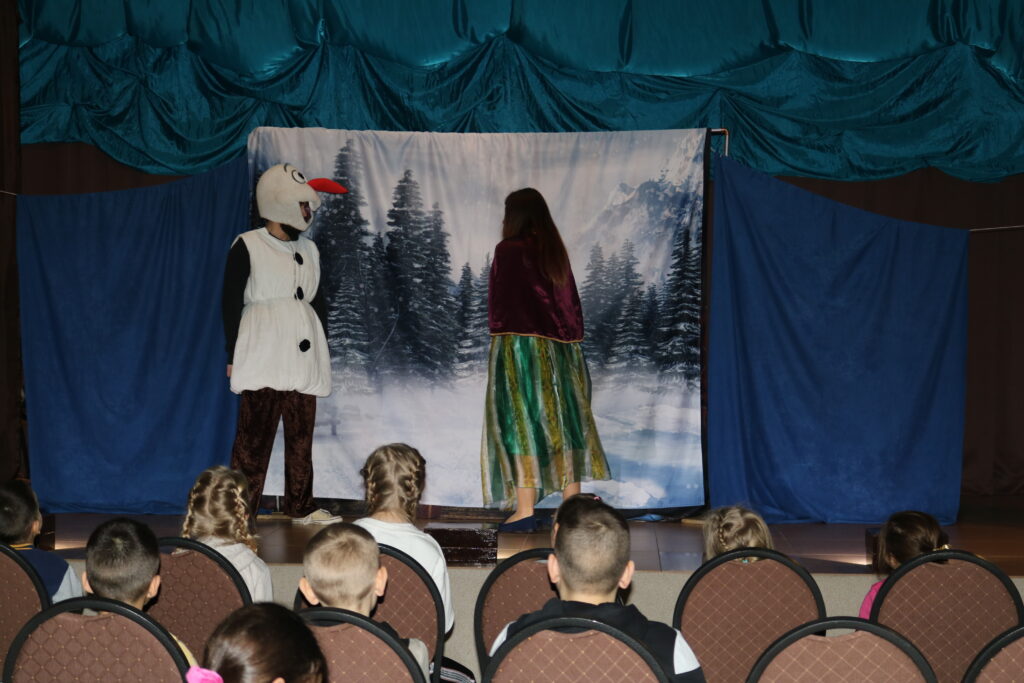 Zdjęcie przedstawia scenę, na której jest dwójka aktorów . Bałwan Olaf i Anna, bohaterowie  bajki Kraina Lodu. Sceneria jest w niebieskich odcieniach, a na środku  baner  z leśnym zimowym krajobrazem. Z przodu sceny na krzesełkach siedzą dzieciaki .