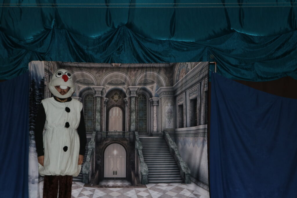 Zdjęcie przedstawia aktora przebranego za bałwanka Olafa, na tle wnętrza zamku.