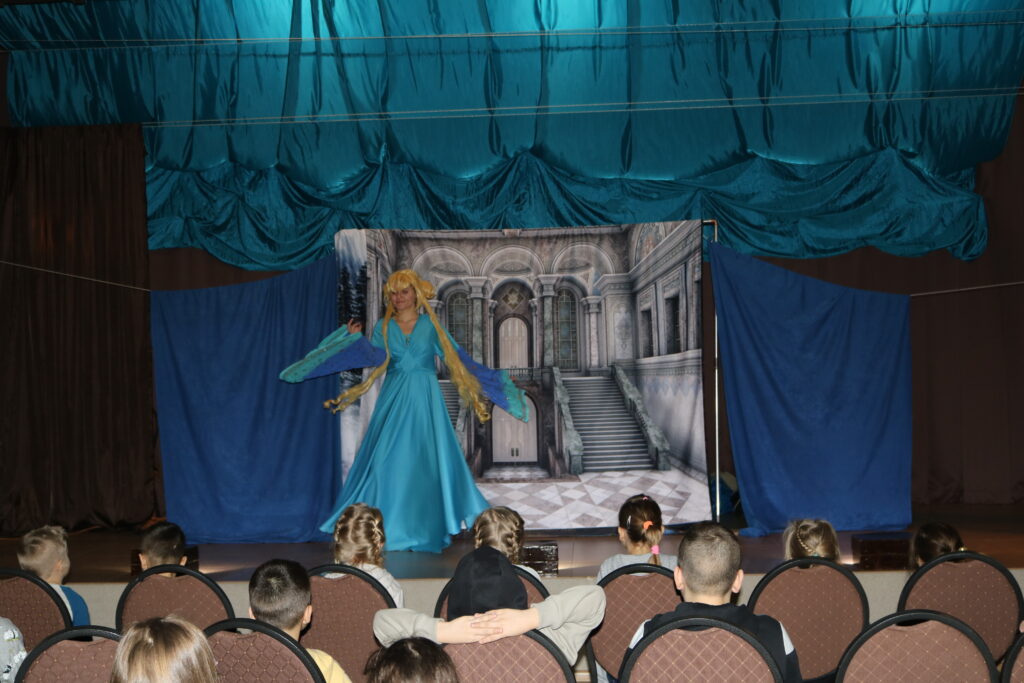Dzieciaki przed sceną , siedzące na krzesełkach wpatrzone w księżniczkę Elzę, która stoi na tle komnaty zamku.
